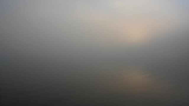 Minimal foggy morning scene at a lake as the sun rises.  Small waves can be seen in the water and the water reflects the colors of the fog and sun.
