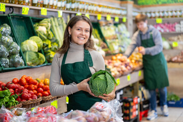 Positive young female seller holding cabbage standing by counter in vegetable market