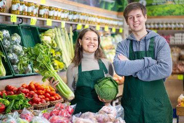 Happy young male and female sellers posing in grocery market with vegetables in hands