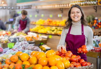 Positive young female seller holding tomatoes standing by counter in vegetable market