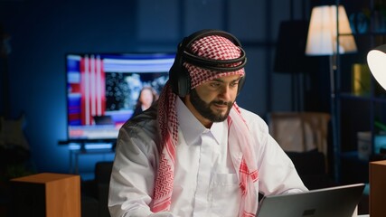 Arab worker with wireless headphones enthusiastic about working from home office. Cheerful Middle Eastern businessman doing online research while listening to music to make time pass faster