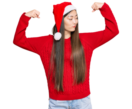 Young chinese woman wearing christmas hat showing arms muscles smiling proud. fitness concept.