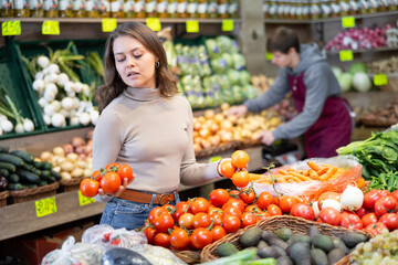 Positive young girl choosing tomatoes standing at counter in large vegetable market