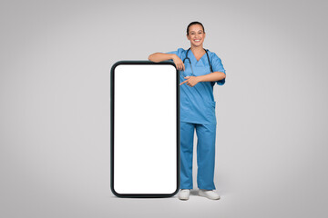 Cheerful lady doctor nurse in blue uniform near big phone pointing at blank screen, isolated on grey studio background