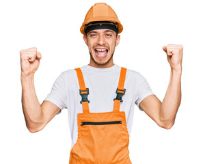 Hispanic young man wearing handyman uniform and safety hardhat screaming proud, celebrating victory and success very excited with raised arms