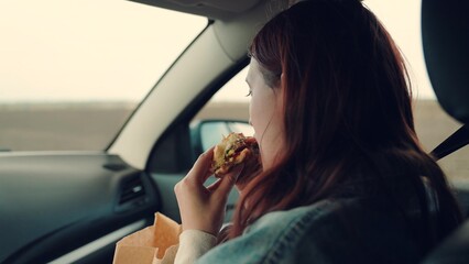 Girl passenger eats burger in car. Delicious food bun with cutlet. Young hungry woman eats roll with meatball while sitting in car. Offspring travels, having snack on road without leaving car.Tourist - obrazy, fototapety, plakaty