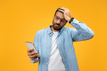 A perplexed man in a blue shirt holds his smartphone and looks at it with a confused expression