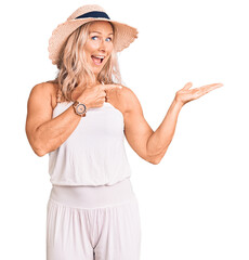 Middle age fit blonde woman wearing summer hat amazed and smiling to the camera while presenting with hand and pointing with finger.