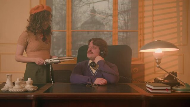 Retro style, young female subordinate brings documents to her boss talking on phone. Vintage office and clothes, employee girl give papers to her boss at newspaper office to sign.