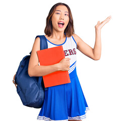 Young beautiful chinese girl wearing cheerleader uniform and student backpack holding binder celebrating victory with happy smile and winner expression with raised hands