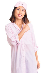 Young beautiful chinese girl wearing sleep mask and pajama cheerful with a smile of face pointing with hand and finger up to the side with happy and natural expression on face