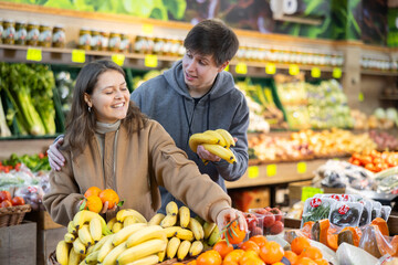 Couple of young woman and young guy choose tangerines and bananas in vegetable shop