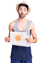Young hispanic man wearing summer hat holding sun draw scared and amazed with open mouth for...