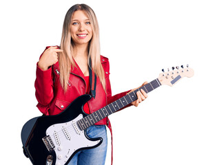 Young beautiful blonde woman playing electric guitar pointing finger to one self smiling happy and...