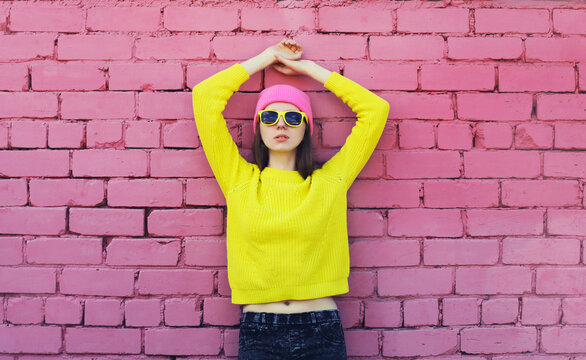 Portrait of stylish young teenager girl wearing vivid colorful clothes, knitted yellow sweater and pink hat on brick wall background