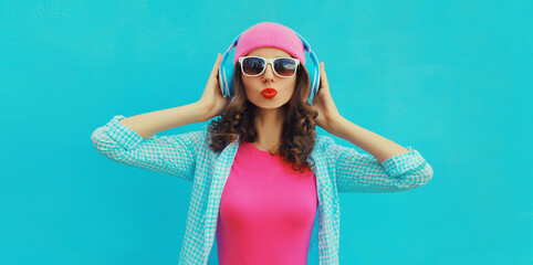 Portrait of stylish modern happy young woman listening to music with headphones wearing colorful pink hat on blue studio background