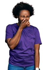 African american woman with afro hair wearing casual purple t shirt smelling something stinky and...
