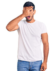 Young latin man wearing casual clothes peeking in shock covering face and eyes with hand, looking through fingers with embarrassed expression.
