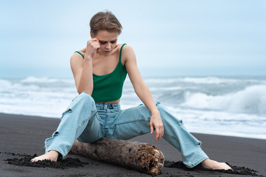 Blonde woman sitting on log with her legs spread and head lowered thoughtfully, looking down on black sandy beach of Pacific Ocean. Millennial woman with short hair wearing blue jeans and green top