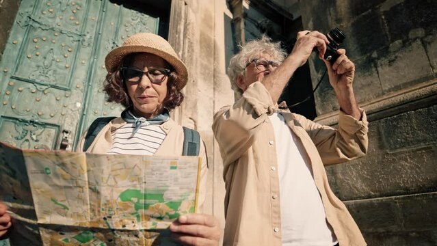 Camera view from below of older Caucasian couple standing in front of large green doors of old church or museum. Woman holding physical map while man taking photos of wonderful scenery.