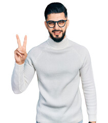 Young arab man with beard wearing elegant turtleneck sweater and glasses showing and pointing up with fingers number two while smiling confident and happy.