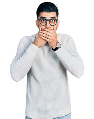 Young arab man with beard wearing elegant turtleneck sweater and glasses shocked covering mouth...
