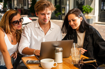 Three young adults sit at a bar table outside for breakfast  in a bright sunny day laughing together while watching contents on a laptop