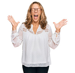 Fototapeta na wymiar Middle age blonde woman wearing casual white shirt and glasses celebrating mad and crazy for success with arms raised and closed eyes screaming excited. winner concept