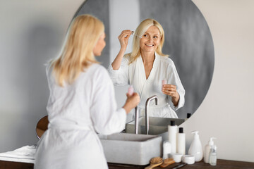 Elegant senior woman in bathrobe applying facial serum near mirror in bathroom