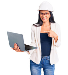 Young beautiful latin girl wearing architect hardhat holding laptop smiling happy pointing with hand and finger