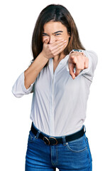 Young caucasian girl wearing casual white shirt laughing at you, pointing finger to the camera with hand over mouth, shame expression
