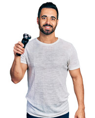 Young hispanic man with beard holding electric razor machine looking positive and happy standing...