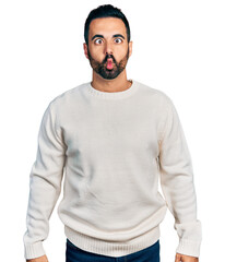 Young hispanic man with beard wearing casual white sweater making fish face with lips, crazy and...