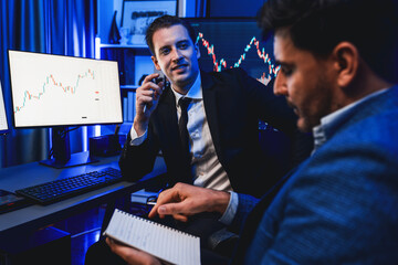 Two stock exchange traders discussing on dynamic investment graph, trending of high profit in currency stock. Showing financial benefit data on monitor screen in neon light at workplace. Sellable.
