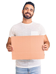 Young hispanic man holding banner cardboard looking positive and happy standing and smiling with a confident smile showing teeth