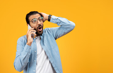 Shocked man in a blue denim shirt talking on a cellphone, holding his head in disbelief or receiving