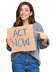 Young brunette woman holding act now banner smiling happy pointing with hand and finger