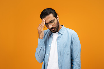 A pensive man in a light blue denim shirt touches his forehead, displaying a gesture of worry - obrazy, fototapety, plakaty