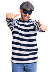 Young handsome man wearing burglar mask approving doing positive gesture with hand, thumbs up...