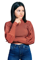 Young brunette woman with blue eyes wearing elegant sweater with hand on chin thinking about question, pensive expression. smiling with thoughtful face. doubt concept.