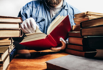 Photo of antiquarian man in a blue shirt and white protective gloves holding and reading red antique book and sitting by a table with books pile.