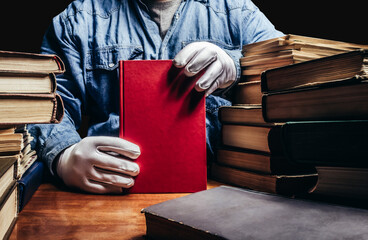 Photo of antiquarian man in a blue shirt and white protective gloves holding a red book and sitting...