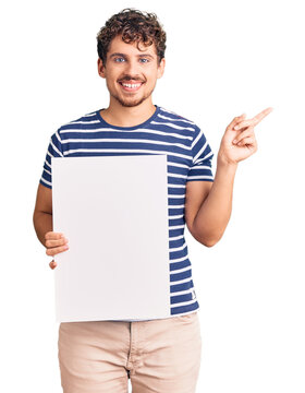 Young handsome man with curly hair holding blank empty banner smiling happy pointing with hand and finger to the side