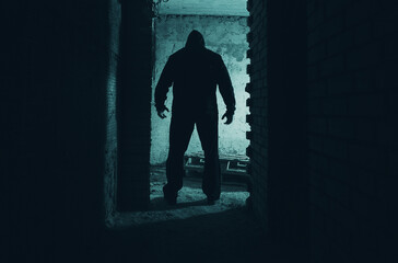 Horror photo of a man in black hood, stalker or killer standing in a doorway in the dark abandoned...