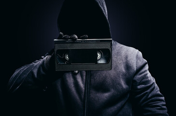 Photo of scary shaded hooded man showing and holding video cassette tape on dark background.