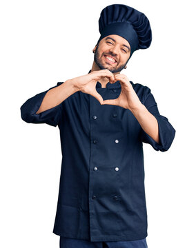 Young hispanic man wearing cooker uniform smiling in love doing heart symbol shape with hands. romantic concept.