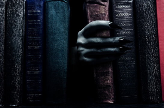 Photo of scary haunted female witch hand with claws reaching out between books.