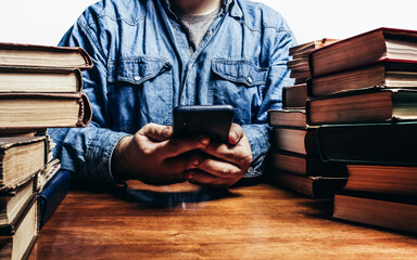 Photo of male person in denim shirt holding smartphone and sitting by the table with old book stacks, selling antiquities or audio books concept.