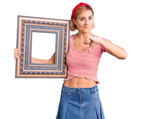 Young blonde woman holding empty frame with angry face, negative sign showing dislike with thumbs down, rejection concept