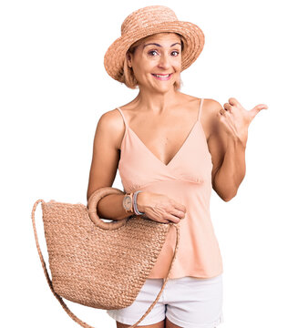 Young blonde woman wearing summer hat holding bag pointing thumb up to the side smiling happy with open mouth